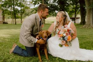 Bride and groom kneel in grass with their brown dog wearing a floral collar at an outdoor wedding.