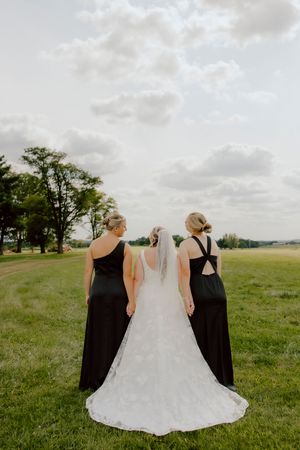 Bride with two bridesmaids in black dresses walking through an open green field at sunset.