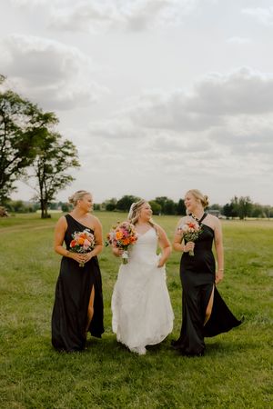 Bride in white gown walks with two bridesmaids in black dresses holding colorful bouquets in a green field.