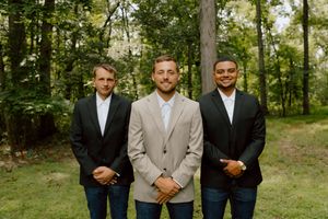 Groom poses with two groomsmen in dark blazers and jeans in a wooded outdoor setting.