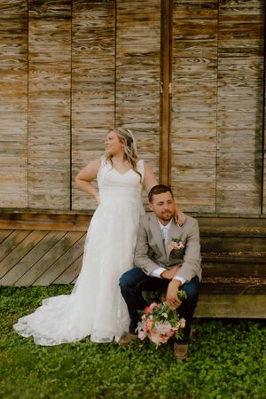 Bride stands behind seated groom holding a bouquet in front of a rustic wooden barn wall.
