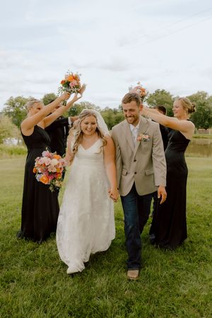 Smiling bride and groom walk hand-in-hand as bridesmaids in black dresses raise bouquets overhead outdoors.