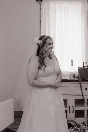 Elegant bride in lace wedding gown and veil smiles softly in a black and white portrait by a window.