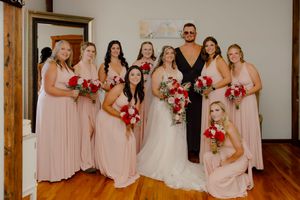 Bride poses with bridesmaids in blush gowns holding red and white bouquets inside a rustic venue.