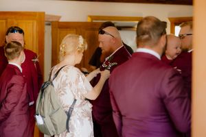 Wedding guest pins a boutonniere on a groomsman in a burgundy suit before the ceremony.