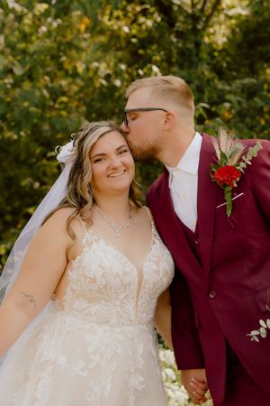 Groom in burgundy suit kissing bride on cheek, bride smiling in lace wedding gown outdoors.