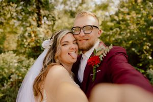 Bride and groom taking a fun selfie outdoors, bride sticking out tongue, groom in burgundy suit.