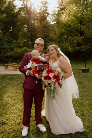 Bride and groom in red suit holding baby with red floral bouquet outdoors at wedding ceremony.