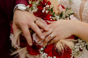 Close-up of bride and groom wedding rings on hands over red floral bouquet.