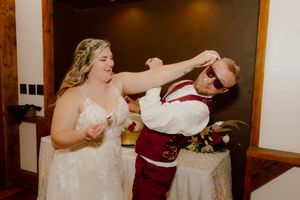 Laughing bride and groom in burgundy suit playfully cut their wedding cake at the reception.