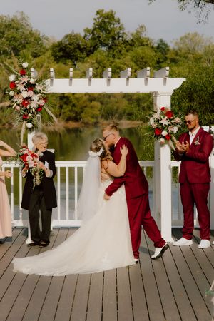 Groom dips bride for a romantic first kiss under a white pergola at a lakeside wedding ceremony.