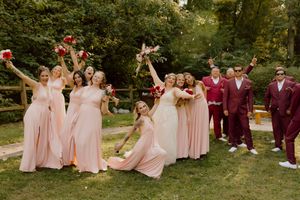 Wedding party celebrating outdoors, bridesmaids in blush pink gowns and groomsmen in burgundy suits raising bouquets.
