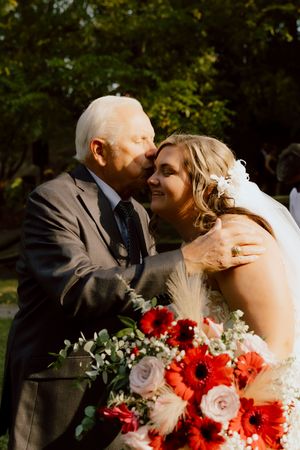 Elderly man in gray suit kissing bride on cheek, bride holding red and pink floral bouquet outdoors.