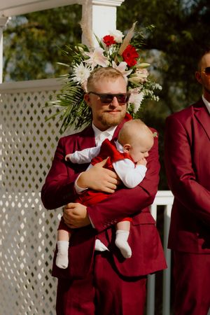 Groomsman in burgundy suit holds an adorable baby dressed in red at an outdoor wedding ceremony.