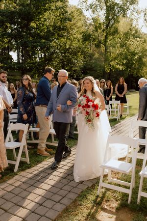 Bride walks down the aisle with her father holding a vibrant red and white bouquet at outdoor wedding.