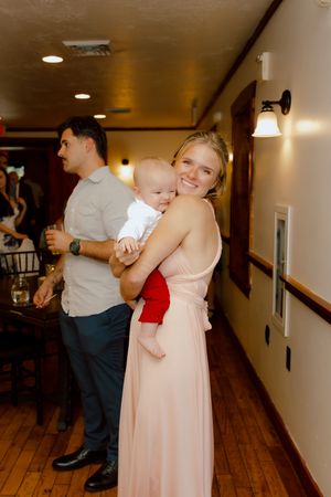 Woman in blush pink gown holds a baby in red pants at wedding reception.