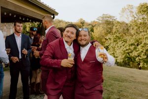 Two groomsmen in burgundy suits smile and pose together at outdoor wedding cocktail hour.