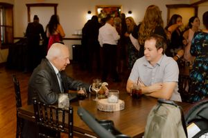 Two male wedding guests share conversation seated at a wooden table during reception.