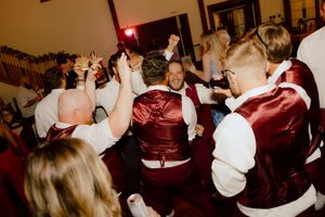 Groomsmen in burgundy satin vests dancing energetically at a lively wedding reception.