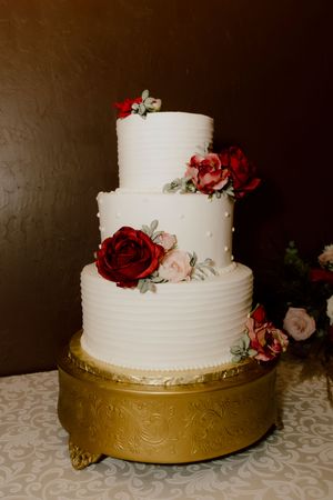 Three-tier white wedding cake with red and pink roses on gold ornate cake stand.