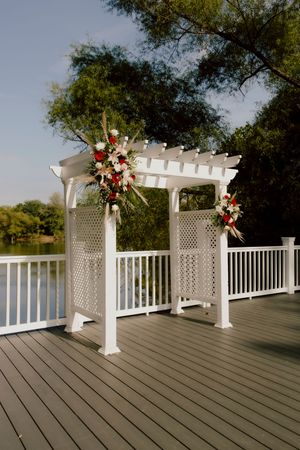 White lattice wedding arch decorated with red and pink floral arrangements on outdoor deck.