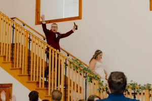 Groom celebrates with raised arms on a staircase decorated with greenery as bride descends beside him.