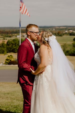 Newlyweds share a first kiss in front of an American flag with a beautiful rural landscape behind them.