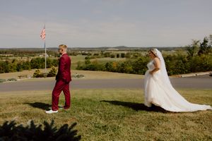 Groom in red suit sees bride in white wedding gown for the first time on a scenic hilltop.