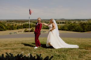 Bride in white gown and groom in red suit hold hands on a hilltop with American flag backdrop.