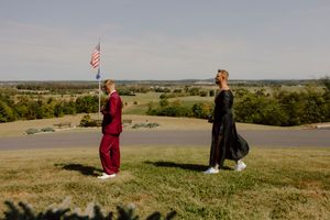 Couple in formal attire standing apart on a hilltop with an American flag and scenic countryside views.