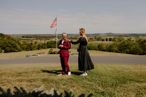 Couple in red suit and black dress share a moment on a scenic hilltop with an American flag nearby.
