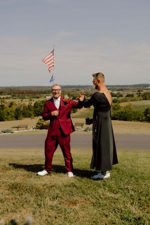 Groom in red suit holds hands with partner in black dress on a hilltop with American flag backdrop.