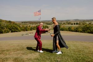 Couple dancing playfully on a hilltop with an American flag and lush green landscape behind them.