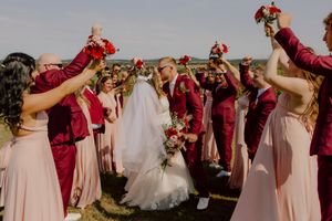 Bride and groom sharing a kiss surrounded by wedding party raising red bouquets in celebration.