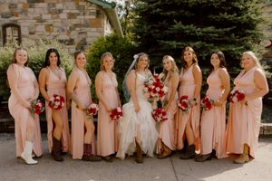 Bride with eight bridesmaids in blush pink gowns holding red bouquets posing outdoors near stone building.