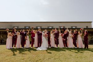 Large wedding party cheering outside barn venue, bridesmaids in blush pink and groomsmen in burgundy suits.