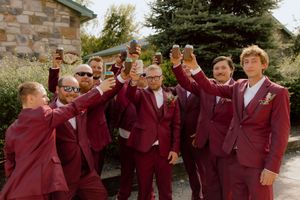 Groomsmen in burgundy suits raising drinks in a toast outdoors on a sunny wedding day.