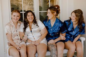 Four bridesmaids in satin pajamas sitting together smiling on outdoor bench before wedding.