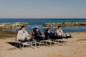 Four groomsmen in suits relaxing on beach lounge chairs by rocky ocean shoreline.