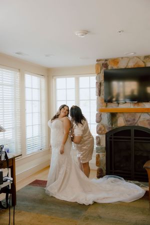 Bride in fitted lace wedding gown being helped by bridesmaid near stone fireplace indoors.