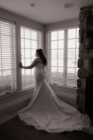 Black and white silhouette of bride in lace gown with train standing by window.