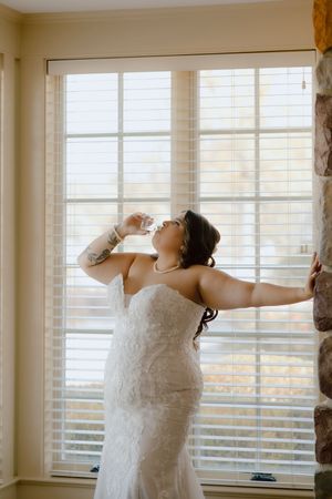 Bride in strapless lace gown drinking from flask with arms outstretched near bright window.