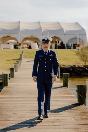 Coast Guard groom in dress uniform walks along wooden dock with white wedding tent in background.