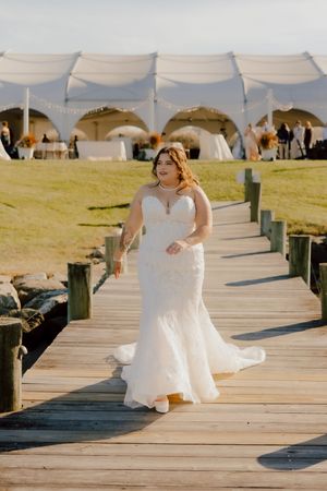 Bride in strapless mermaid wedding gown walks along waterfront dock with white reception tent behind her.