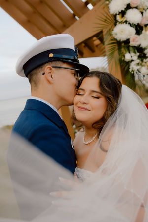 Military groom tenderly kisses bride on cheek as she smiles under floral arch.
