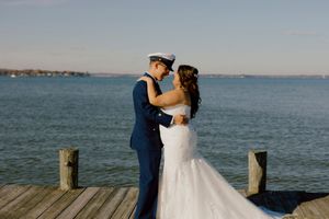 Bride and groom embrace on waterfront dock, groom in Coast Guard uniform and bride in mermaid gown.