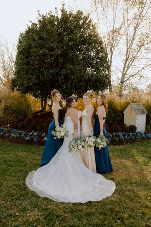 Bride with three bridesmaids in blue and blush gowns holding white bouquets in garden setting.