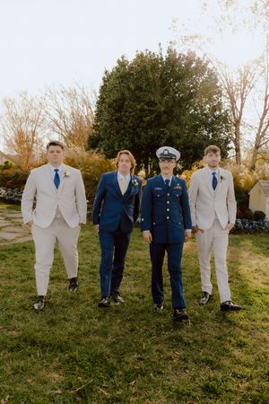 Groom in Coast Guard dress uniform poses with three groomsmen in tan suits in outdoor garden setting.