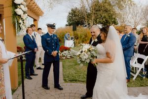 Military officer in dress uniform stands at outdoor wedding ceremony as bride walks down aisle.