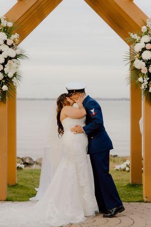 Bride and groom share first kiss under wooden arch with white floral arrangements by waterfront.